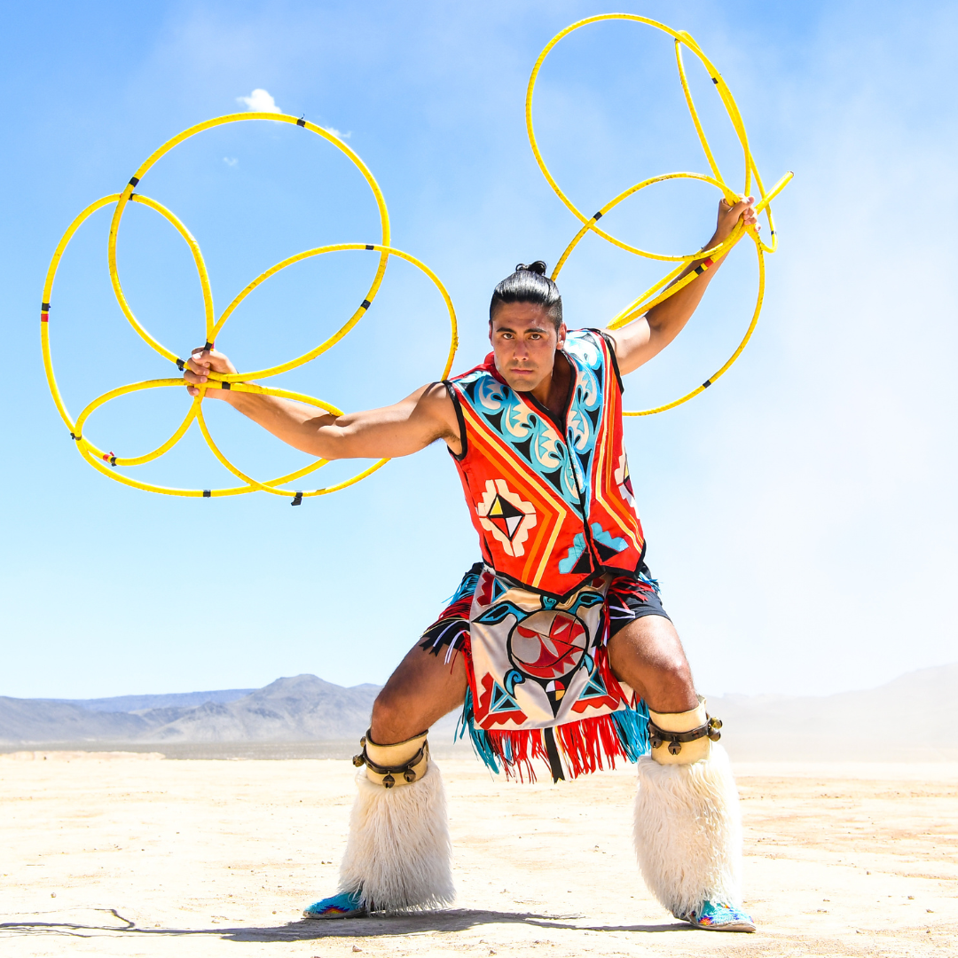 photograph of Eric Hernandez in traditional Lumbee Hoop Dancing regalia. The hoops are bright yellow contrasted with a large, almost infinite blue sky in the desert.