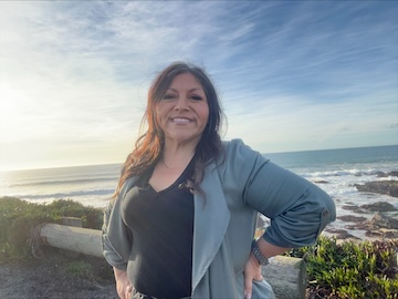 Brijit smiling at the camera with the ocean and bright blue sky in the background